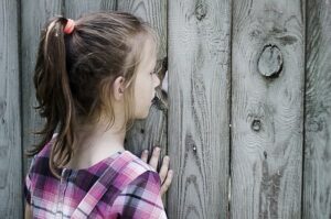 Young girl looking through a hole in the wooden fence